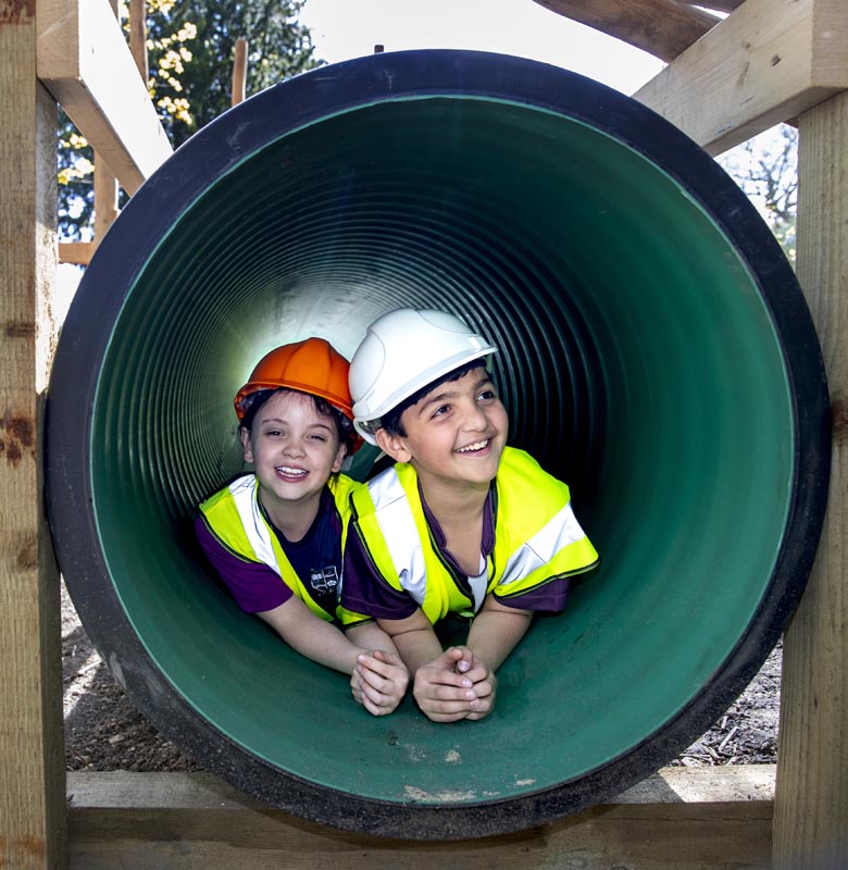Two children wearing safety vests and hard hats inside a large green pipe