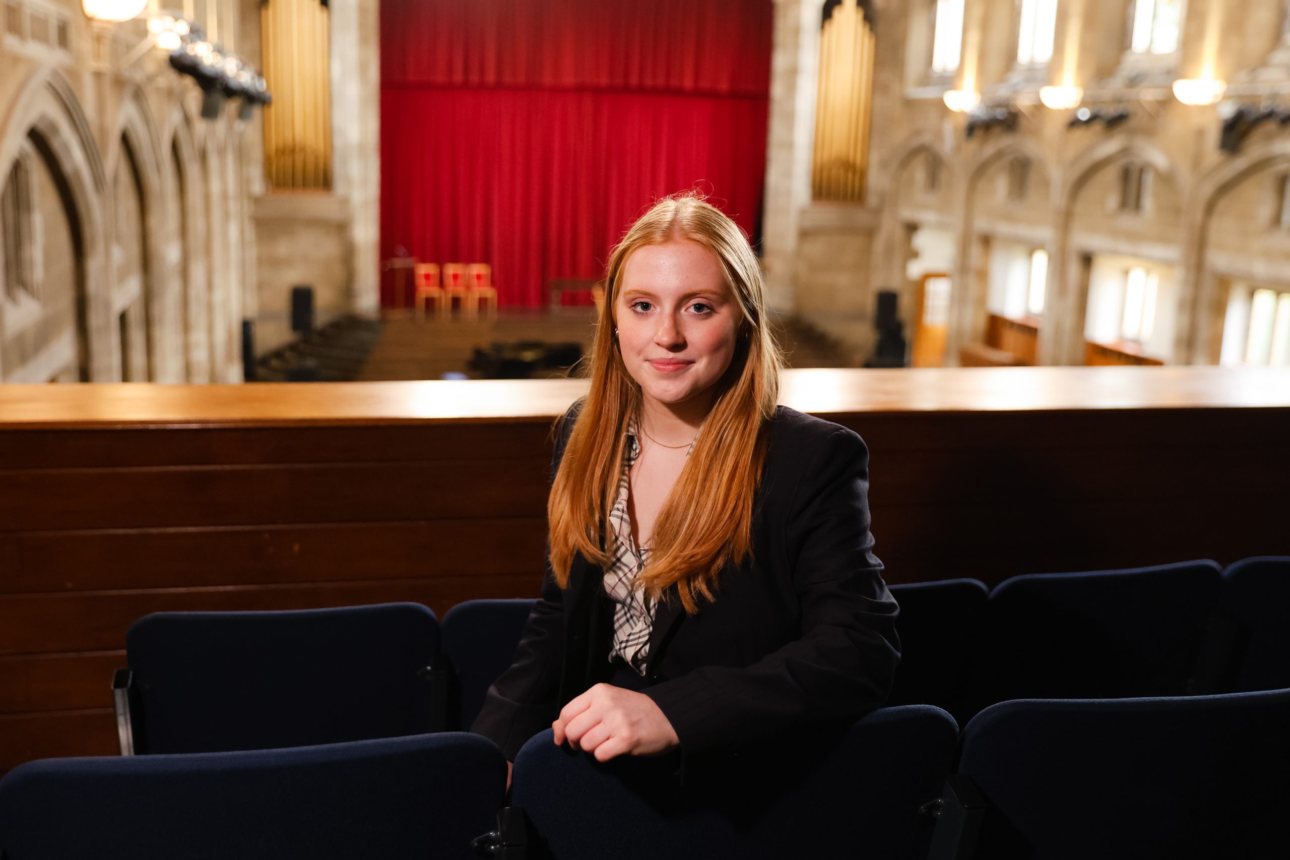 A female student sitting in a hall against a red curtain