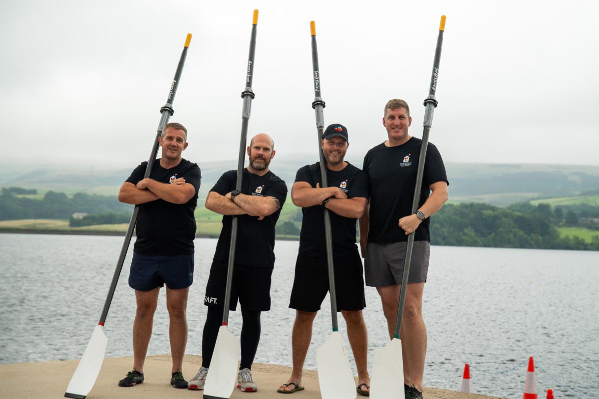 Four men stood with rowing oars on a pier. Wearing black t-shirts with the Ronald McDonald House Charities UK logo on.